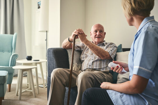 Senior sitzt zuhause auf dem Sofa und wird von einer Betreuerin liebevoll betreut